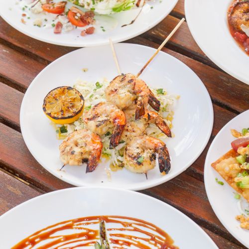 Plates of grilled shrimp and chicken with lemon, cabbage salad, and roasted sides on a wooden table.