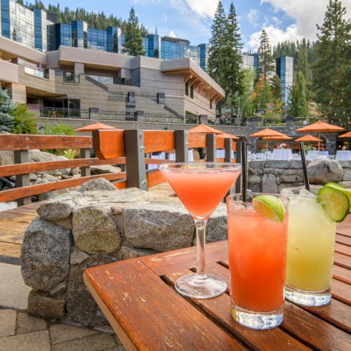 A sunny outdoor patio with orange and green cocktails on a wooden table, pine trees, and an alpine hotel courtyard behind.