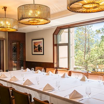 A banquet room set with neatly folded napkins, wine glasses, and chairs under warm chandeliers beside large windows.