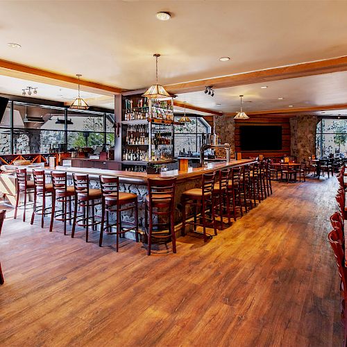 A restaurant with a wooden interior, bar counter with stools, tables with chairs, hanging lights, and large windows, creating a cozy atmosphere.