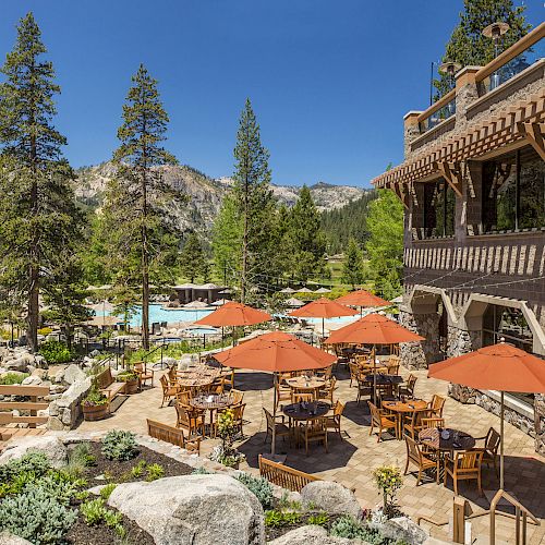 An outdoor dining area with orange umbrellas and wooden tables, situated next to a building named Sandy's Pub, amidst a forested mountain landscape.