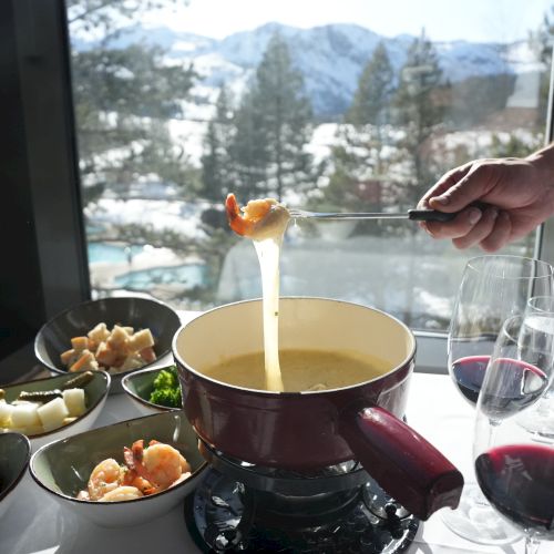 A person dips food into cheese fondue with various ingredients and wine glasses on a table, set against a snowy mountain view through a window.