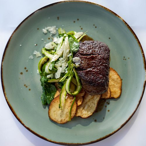 A dish with a piece of grilled meat, sautéed greens, and potato slices served on a light green plate.