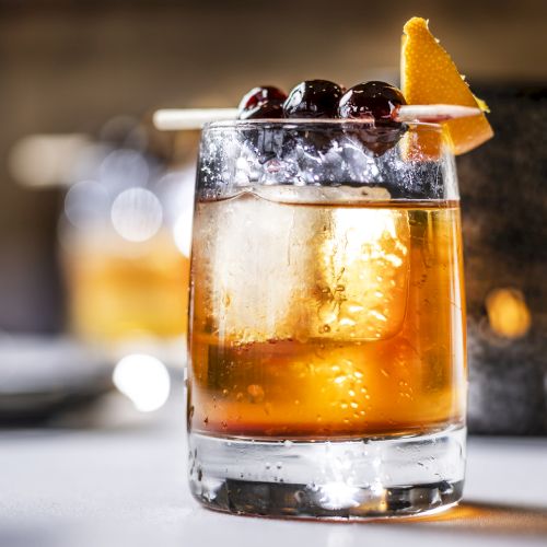 A cocktail in a glass with large ice cubes, garnished with cherries and an orange peel, appears in focus on a blurred background.