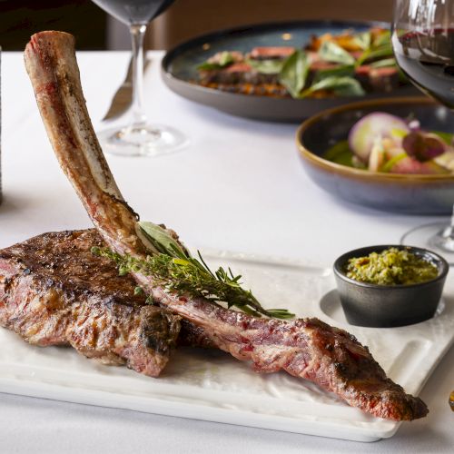 The image shows a large tomahawk steak on a plate, with herbs, a small bowl of sauce, red wine, a lit candle, and additional food dishes in the background.