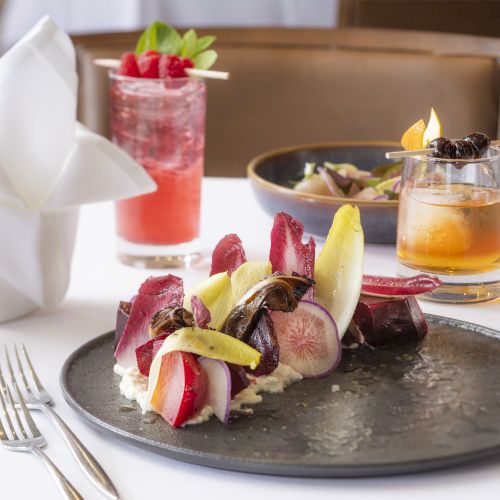 A gourmet dish on a black plate, two colorful cocktails, and a white napkin on a restaurant table.