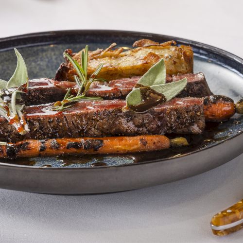 A plate with sliced steak garnished with herbs, accompanied by carrots and roasted potatoes, and a knife placed to the right.