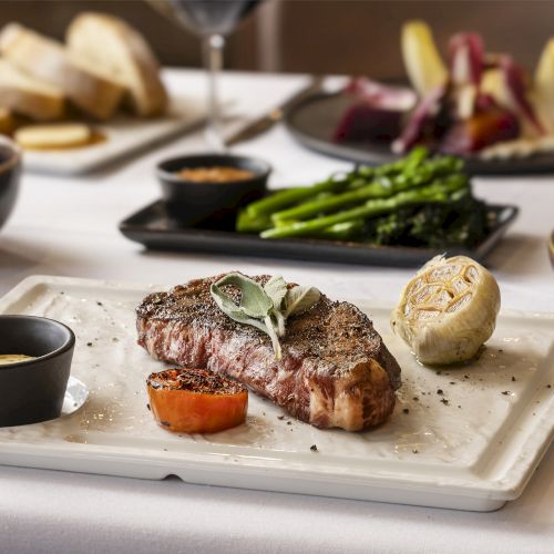 A cooked steak with garnish on a serving plate, accompanied by vegetables, sauce, and a knife. Background shows bread, salad, and other dishes.