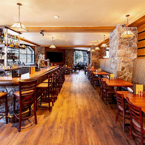 A cozy, well-lit bar and restaurant with wooden chairs and tables, a stone wall, and a stocked bar counter, creating an inviting atmosphere.