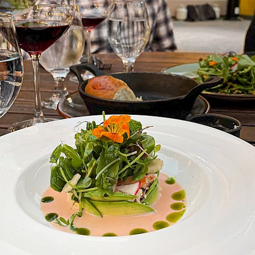 A gourmet dish on a white plate, with wine glasses and a person in the background. Other plates and cutlery are also visible.