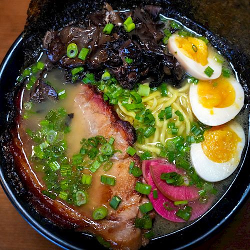 A bowl of ramen with broth, noodles, pork slices, boiled eggs, scallions, mushrooms, and pickled ginger sits on a wooden table.