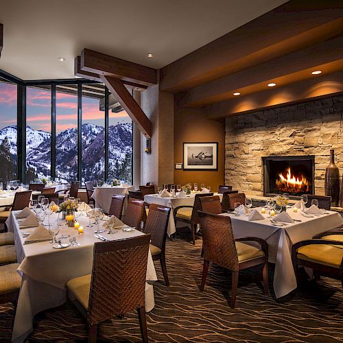 A cozy dining room with elegantly set tables, a fireplace, and large windows showcasing a scenic snowy mountain view during sunset.