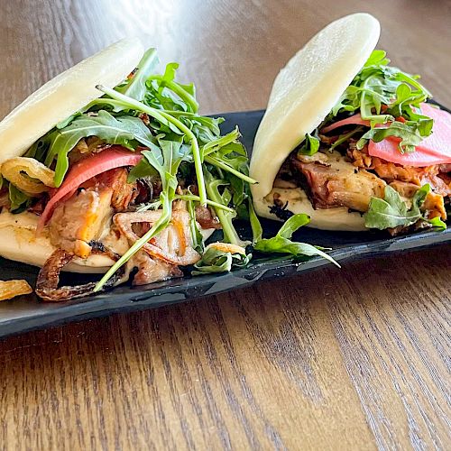 The image shows two steamed bao buns filled with various ingredients, including greens, on a black rectangular plate sitting on a wooden surface.