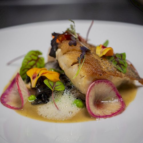 A gourmet dish featuring a piece of fish, colorful vegetables, edible flowers, and sauce, presented artistically on a white plate.