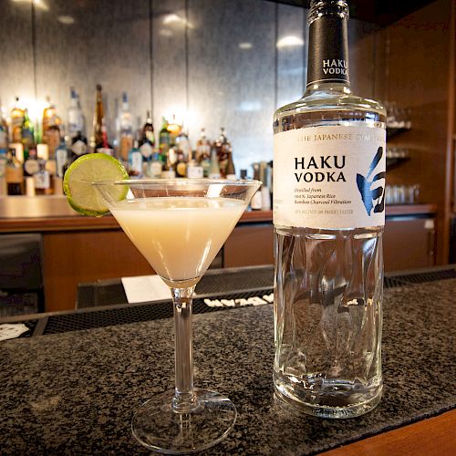 A bottle of Haku Vodka and a cocktail garnished with a lime slice sit on a bar counter, with a stocked bar in the background.