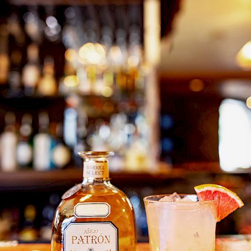 A bottle of Patrón tequila, its cork, and a cocktail garnished with a citrus slice are on a bar counter with a blurred background of shelves with bottles.