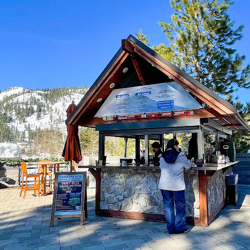 An outdoor kiosk with people standing in front, surrounded by snowy mountains and trees under a clear blue sky.