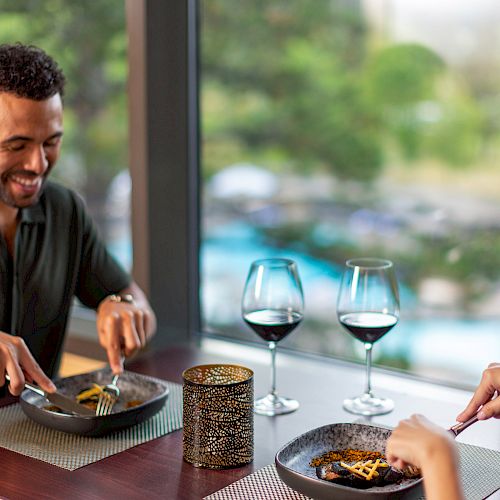 Two people are dining at a restaurant with wine glasses, enjoying a meal together by the window with lush greenery outside.