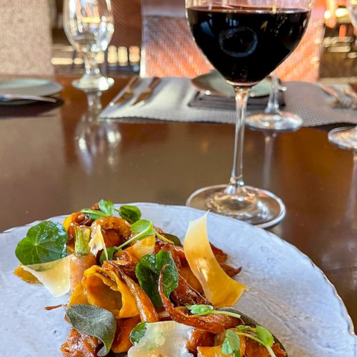 A plate of gourmet food, garnished with greens and vegetables, is in the foreground, with a glass of red wine on a table in the background.