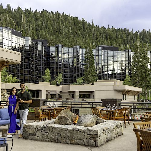 The image shows two people standing by a stone fire pit on an outdoor patio with seating, overlooking modern buildings and a forested hill backdrop.