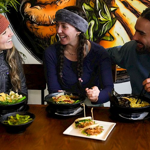 Three friends are enjoying a meal together at a restaurant, smiling and eating from bowls with chopsticks, with several dishes on the table.