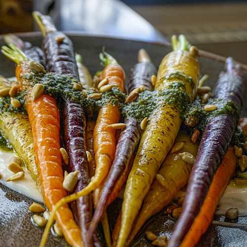 The image shows a plate of vibrant, roasted carrots with various colors, topped with a green herb sauce and scattered with seeds, presented on a creamy sauce.