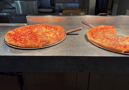 Two large pepperoni pizzas resting on a stainless counter, ready to slice, with a gap between them and a kitchen setup in the background.