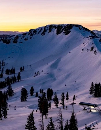 A snowy mountain landscape at sunset with pine trees, a winding trail, and a small building nestled among the slopes, all bathed in warm light.