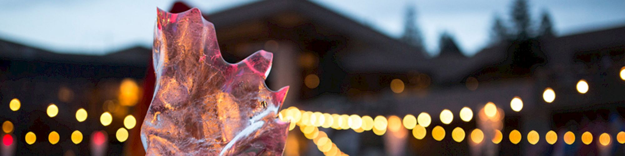 An ice sculpture is illuminated with red lighting in the foreground, with an outdoor event and string lights creating a festive atmosphere in the background.