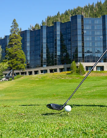 A golfer prepares to tee off on a lush green course with a modern building backdrop; a white golf ball sits near the tee.