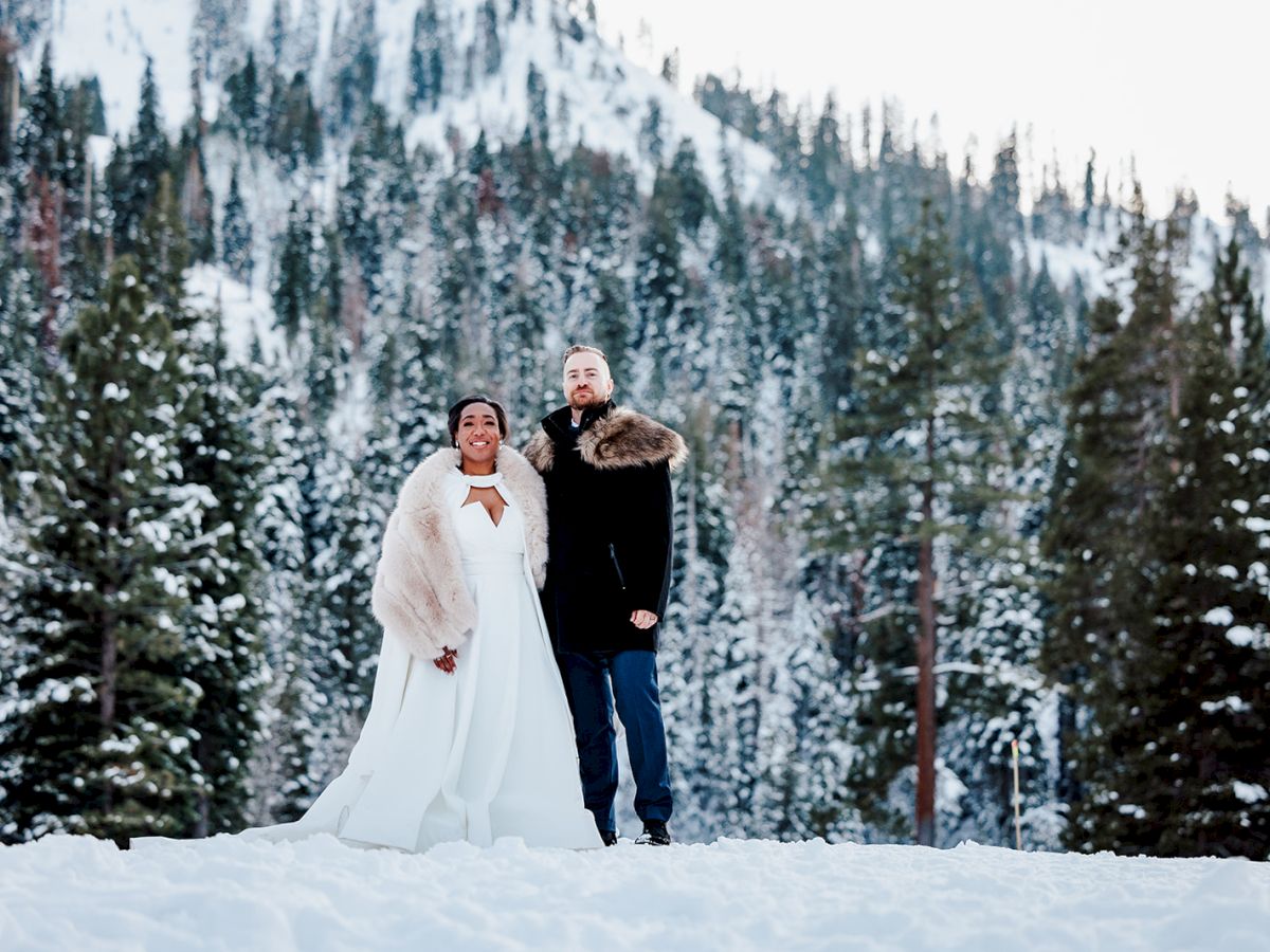 A couple stands in a snowy forest, dressed in wedding attire with a pine forest backdrop and snow-covered ground, posing together.