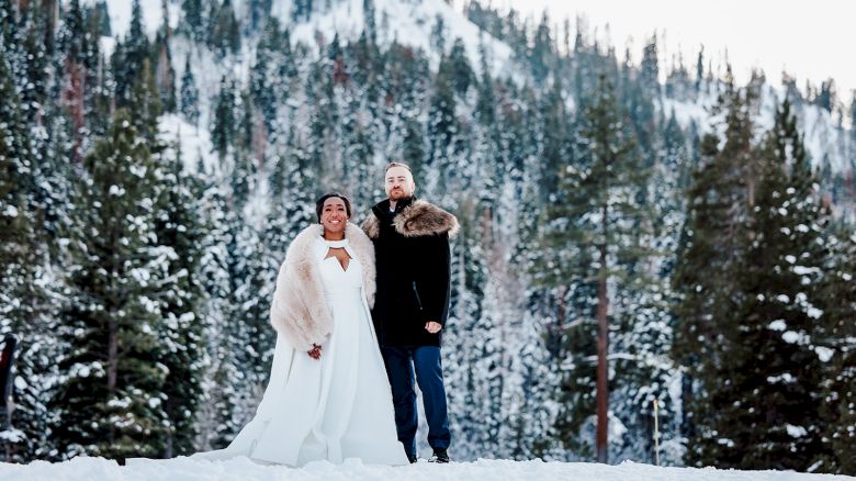 A couple stands in a snowy forest, dressed in wedding attire with a pine forest backdrop and snow-covered ground, posing together.