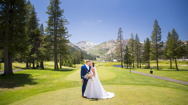 A newlywed couple shares a kiss on a sunny, grassy golf course with mountains in the background, surrounded by tall trees.
