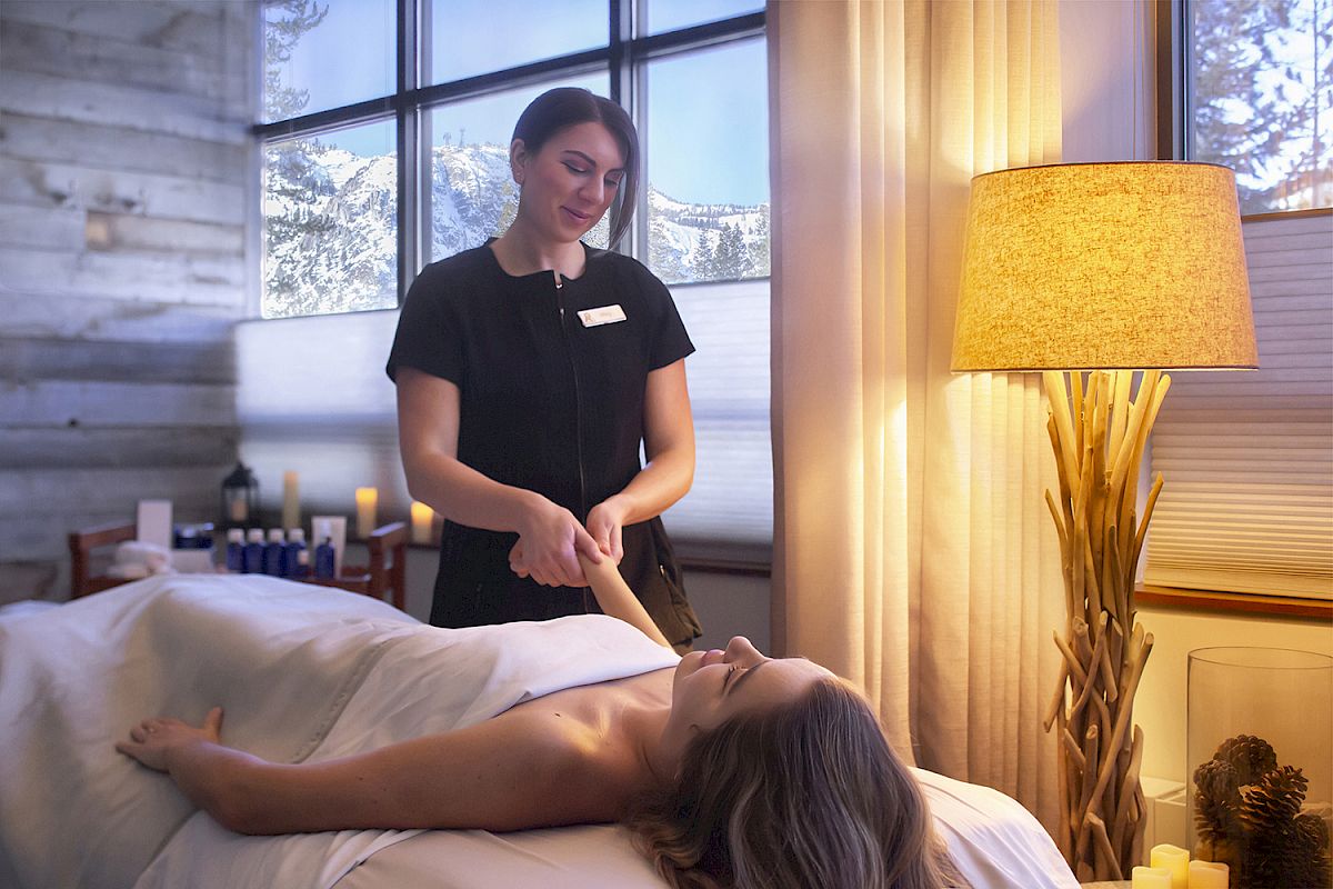 A person is receiving a massage in a cozy room with soft lighting, candles, and a large window view of snow outside.