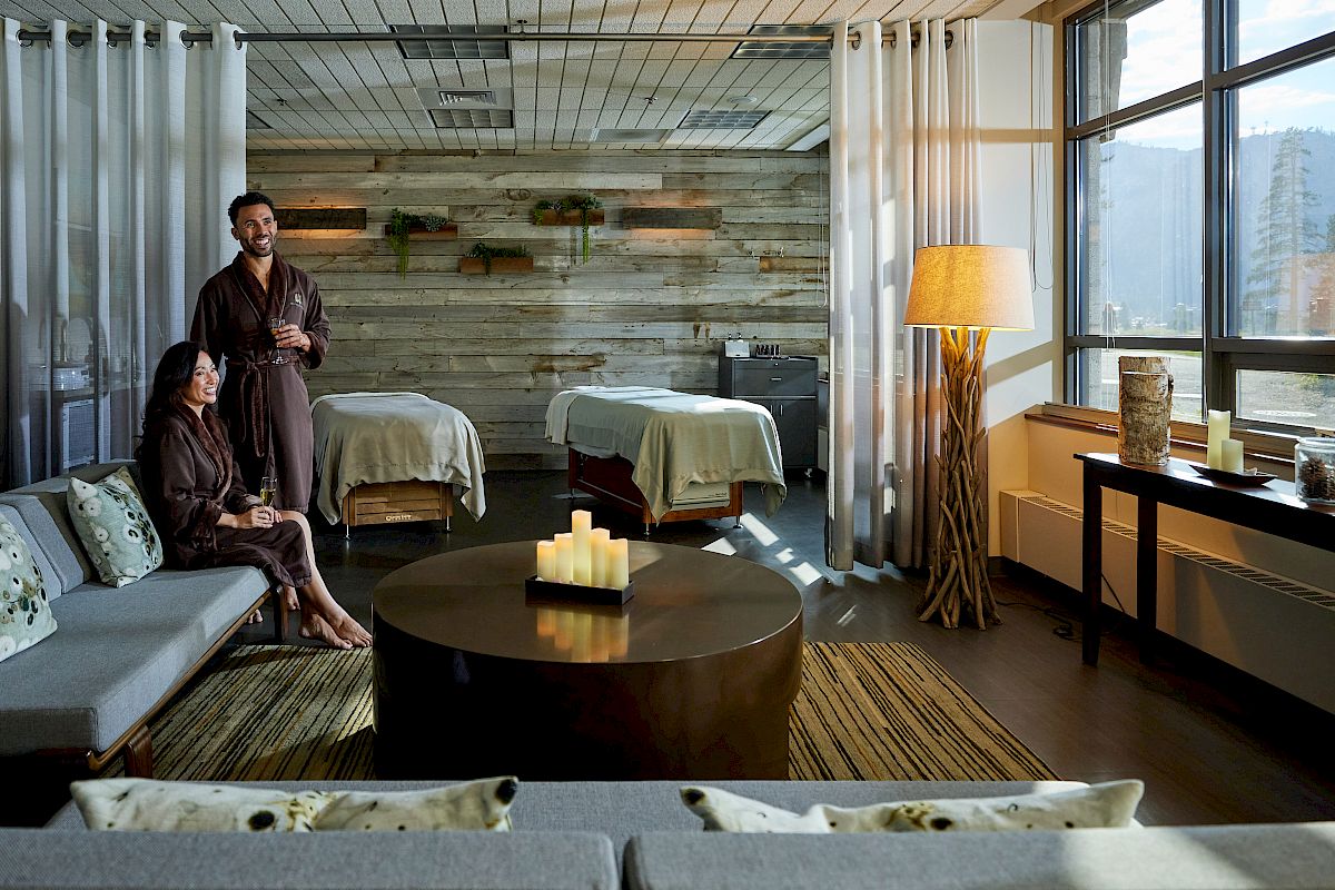 A peaceful spa room with two people in robes, a coffee table with candles, and two massage beds in the background. Warm lighting fills the space.