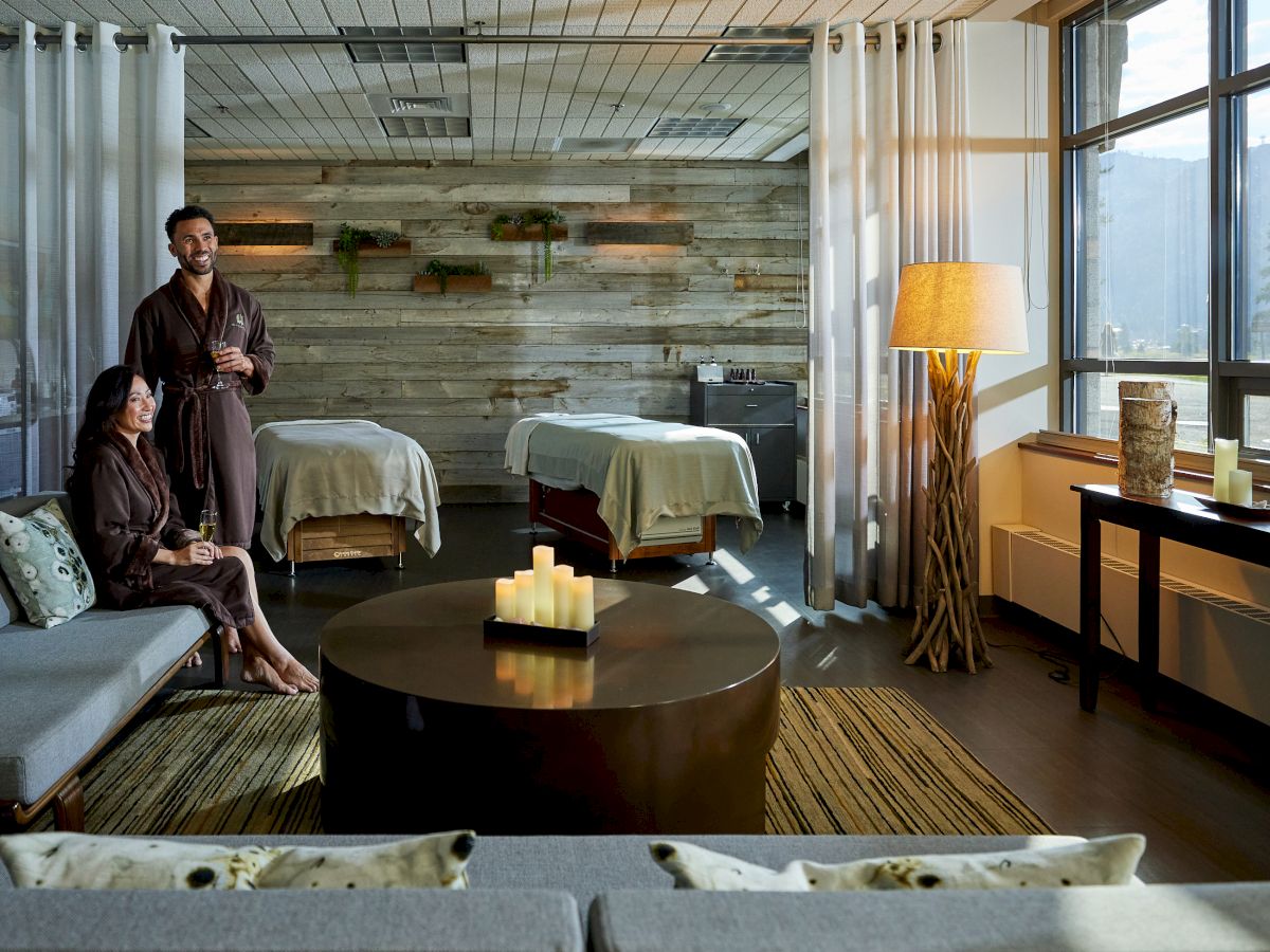 A peaceful spa room with two people in robes, a coffee table with candles, and two massage beds in the background. Warm lighting fills the space.