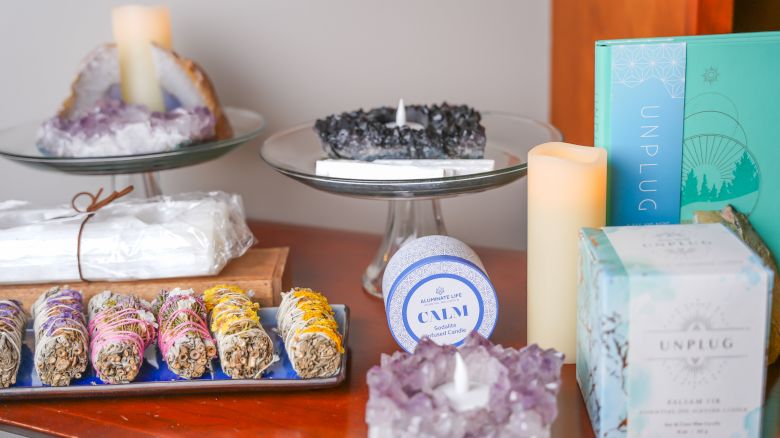 The image shows a display of crystals, candles, sage bundles, and boxed products arranged neatly on a wooden surface.