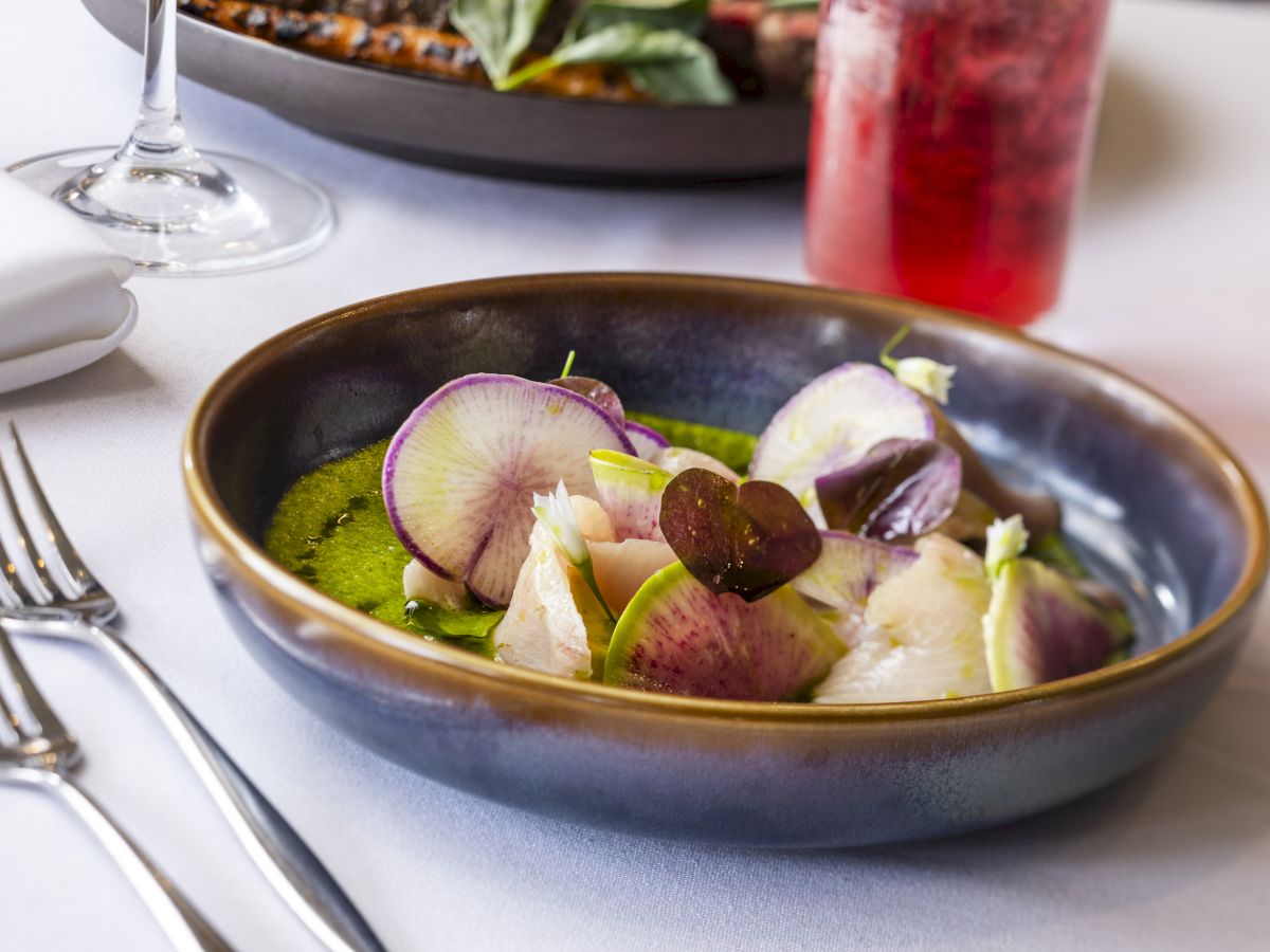 A dish with sliced vegetables and garnish on a green sauce, placed on a table with cutlery, a white napkin, a wine glass, and a red drink.