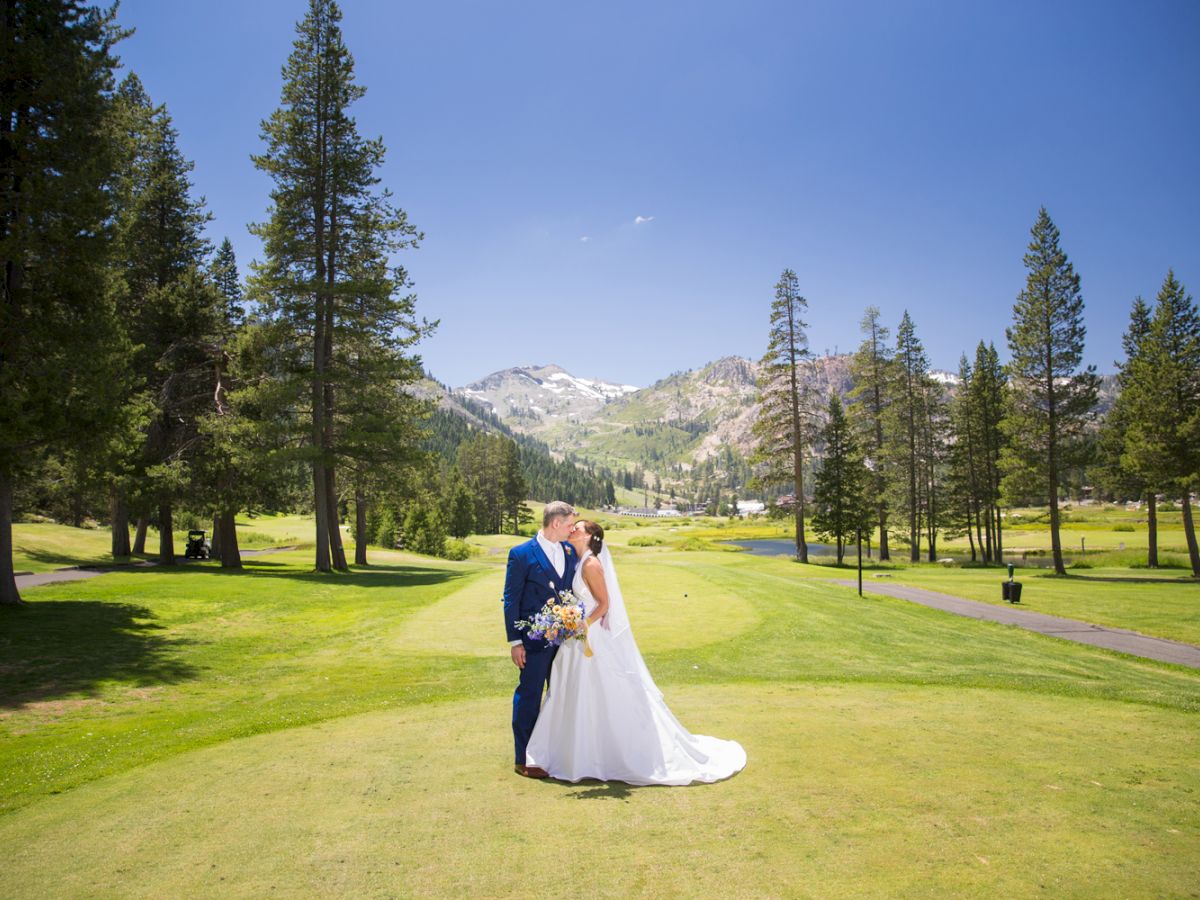 A newlywed couple in formal attire embrace on a sunny green golf course, with tall pines and distant mountains in the background.