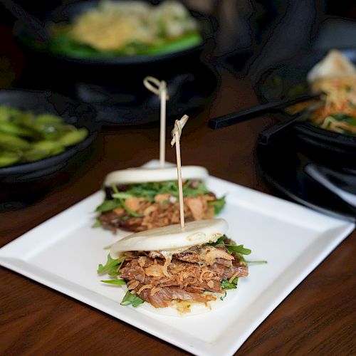 The image shows bao buns with shredded meat and greens on a plate, with edamame and noodle dishes in the background.