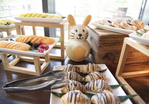 A cafe spread with pastries on skewers, a cute bunny figurine centerpiece, bread rolls, and tongs on a wooden counter. End.