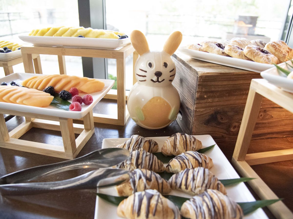 A cozy bakery spread: pastries on wooden racks, glazed donuts, a cute bunny figurine, and a plate of drizzled pastries with tongs nearby.
