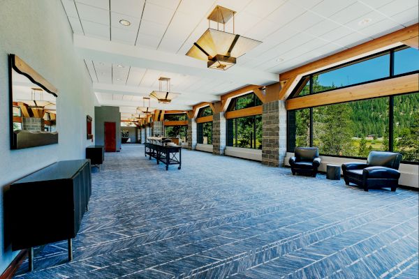 A spacious, modern room with large windows, armchairs, tables, decorative lighting, and mountain views outside. The room has a blue pattern carpet.