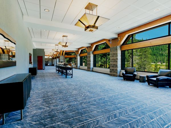 A spacious, modern room with large windows, armchairs, tables, decorative lighting, and mountain views outside. The room has a blue pattern carpet.