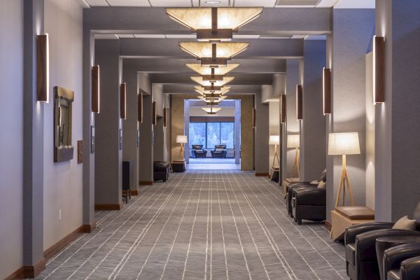 A well-lit, modern hallway with contemporary furnishings, including chairs, lamps, and wall decorations, leading to a seating area by a large window.