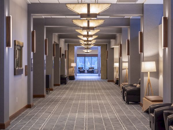 A well-lit, modern hallway with contemporary furnishings, including chairs, lamps, and wall decorations, leading to a seating area by a large window.