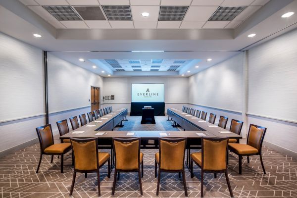 A conference room setup with a U-shaped table, chairs, a projector screen, and a central podium, well-lit with ceiling lights, and carpeted floor.