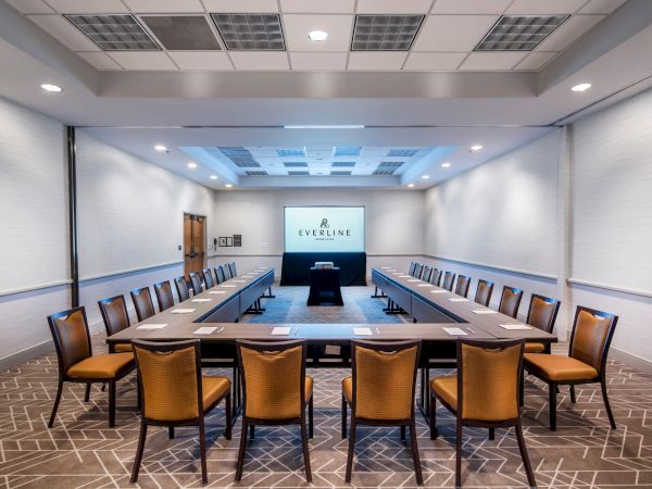 A conference room setup with a U-shaped table, chairs, a projector screen, and a central podium, well-lit with ceiling lights, and carpeted floor.