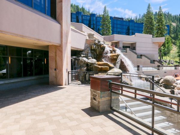 An outdoor stone fountain splashes beside a modern lodge, with pine trees and mountains in the background.