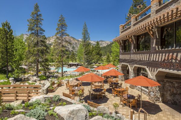 An outdoor dining area with wooden tables and orange umbrellas, set in a lush, green mountain landscape next to a building sign reading 'Sandy's Pub'.